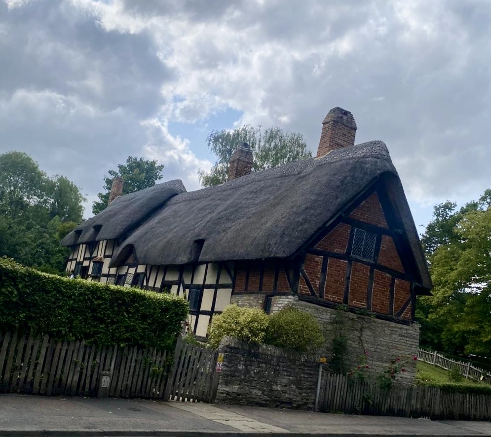 A thatched half-timbered cottage with brick infill panels and multiple chimneys behind a stone wall and wooden fence
